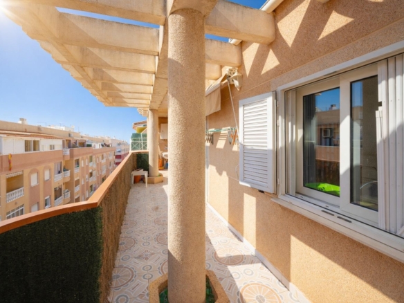 Sunlit balcony with beige walls, a textured column, white shuttered window, and patterned tiled floor.
