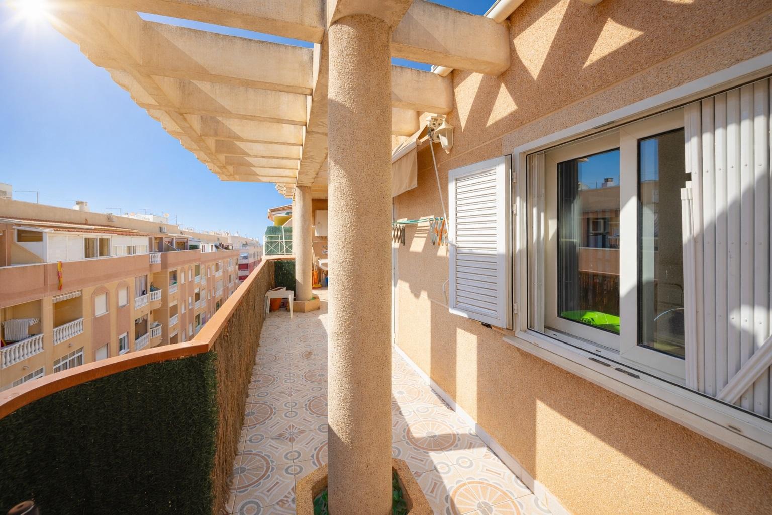 Sunlit balcony with beige walls, a textured column, white shuttered window, and patterned tiled floor.