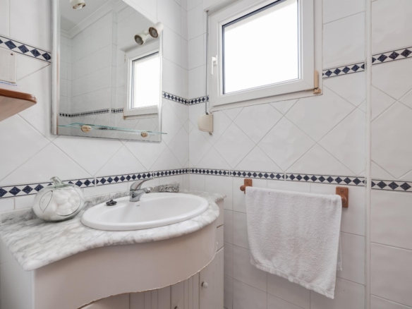 White tiled bathroom with a marble countertop sink, mirror, and a towel hanging on a rack near a window. The blue diamond tile border runs around the walls.