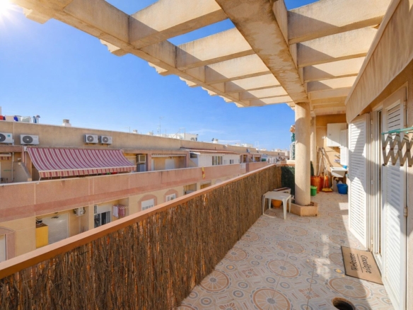 Sunlit apartment balcony with patterned tiled floor, decorative railing, and a pergola overhead; neighboring buildings and clear blue sky in view.