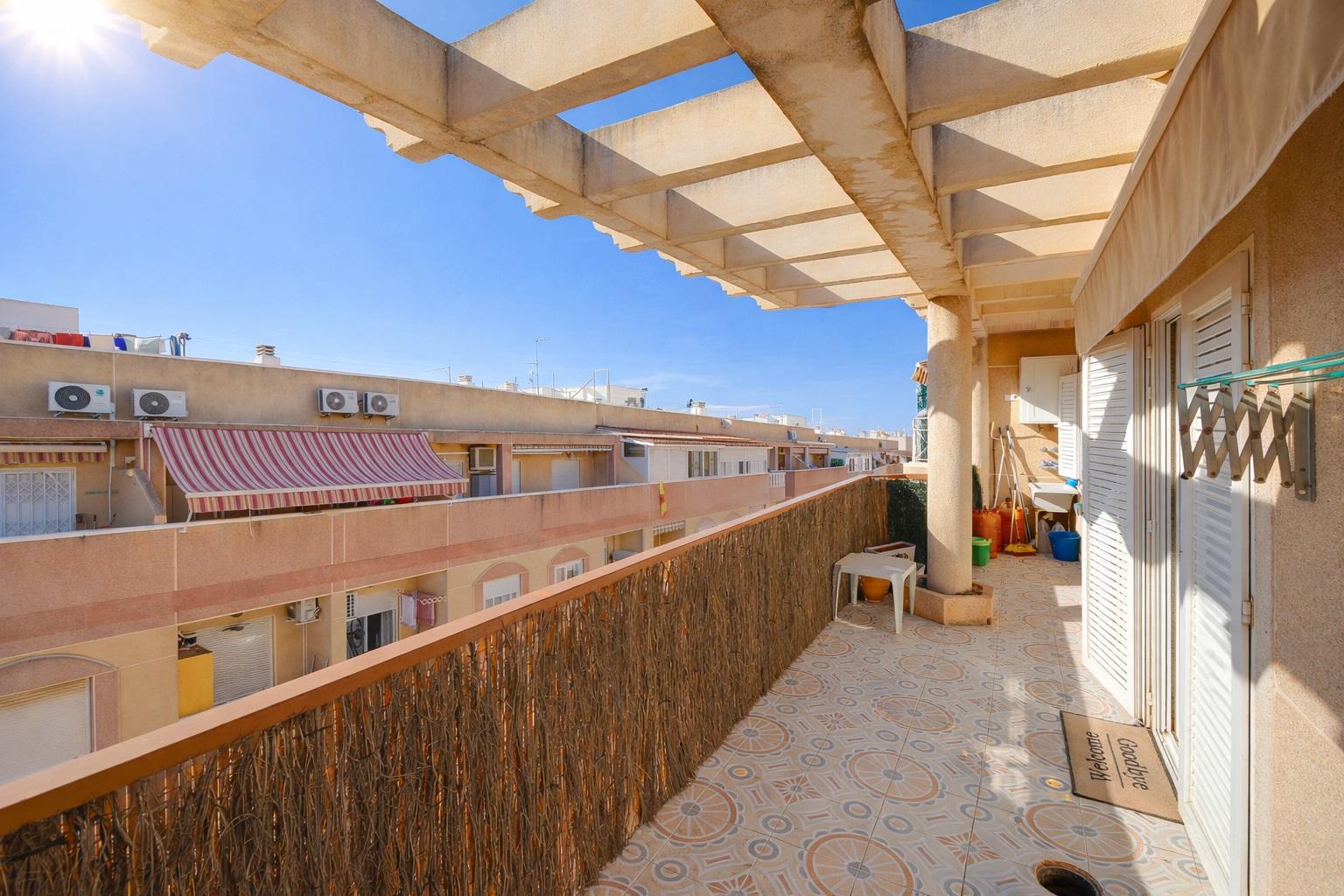 Sunlit apartment balcony with patterned tiled floor, decorative railing, and a pergola overhead; neighboring buildings and clear blue sky in view.