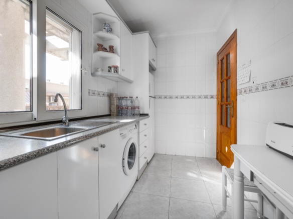Bright white kitchen with a double sink by a large window and a row of white cabinets and a washing machine underneath the counter.