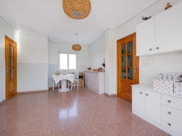 Bright dining/kitchen area with white cabinets, wooden doors, round wicker pendant light, and a small table by a window set for a meal.