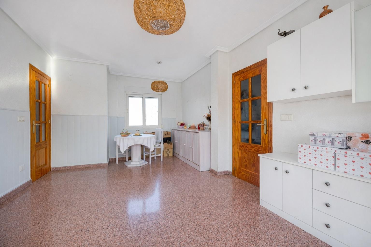 Bright dining/kitchen area with white cabinets, wooden doors, round wicker pendant light, and a small table by a window set for a meal.