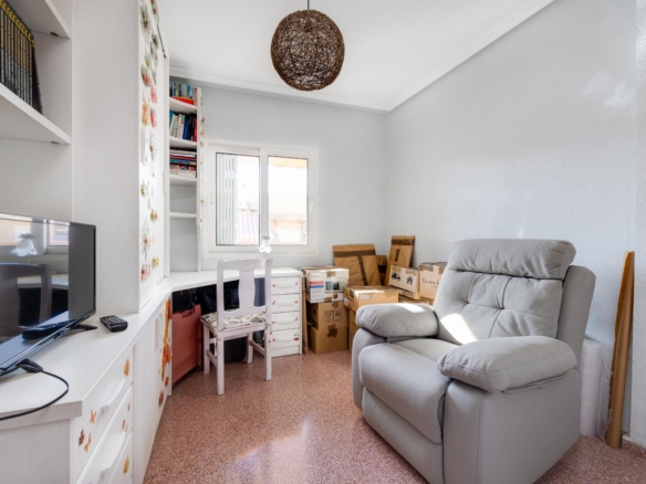 A small living/working space with white shelves, a desk and computer on the left, and stacked cardboard boxes beside a gray recliner by the window.