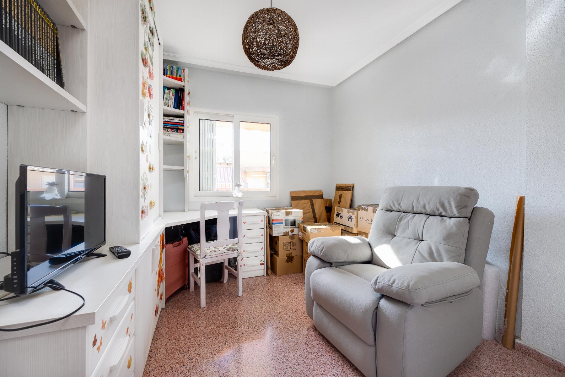 A small living/working space with white shelves, a desk and computer on the left, and stacked cardboard boxes beside a gray recliner by the window.
