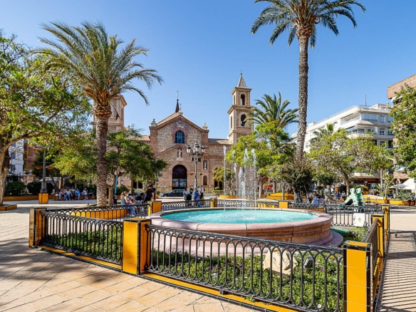 Public square with a circular fountain, yellow-and-black railing, palm trees, and a stone church in the background; people sit and stroll.
