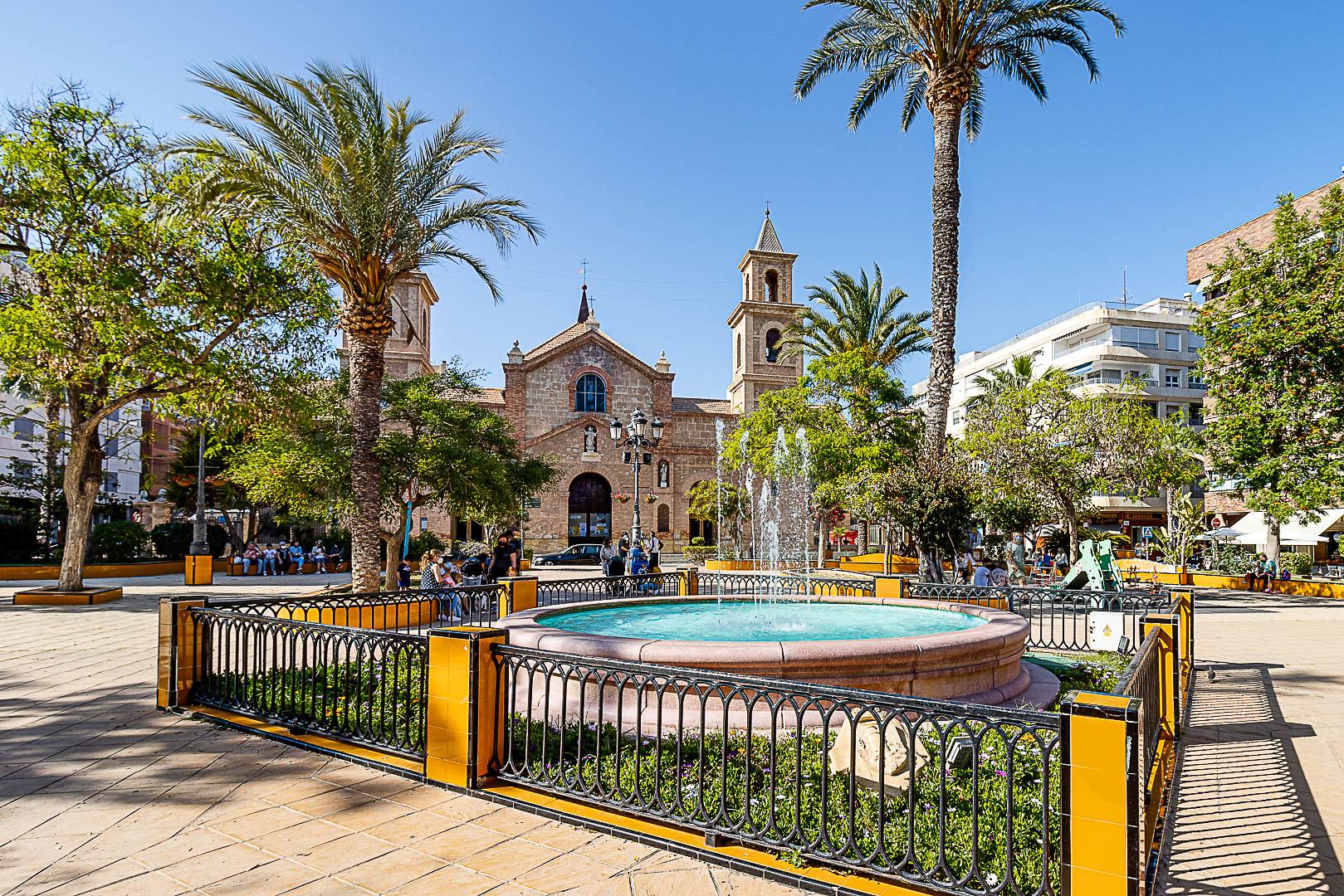 Public square with a circular fountain, yellow-and-black railing, palm trees, and a stone church in the background; people sit and stroll.
