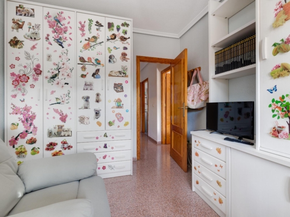 Living room corner with white storage cabinets covered in colorful dog and floral stickers, a gray sofa in foreground, and a TV on a white dresser.