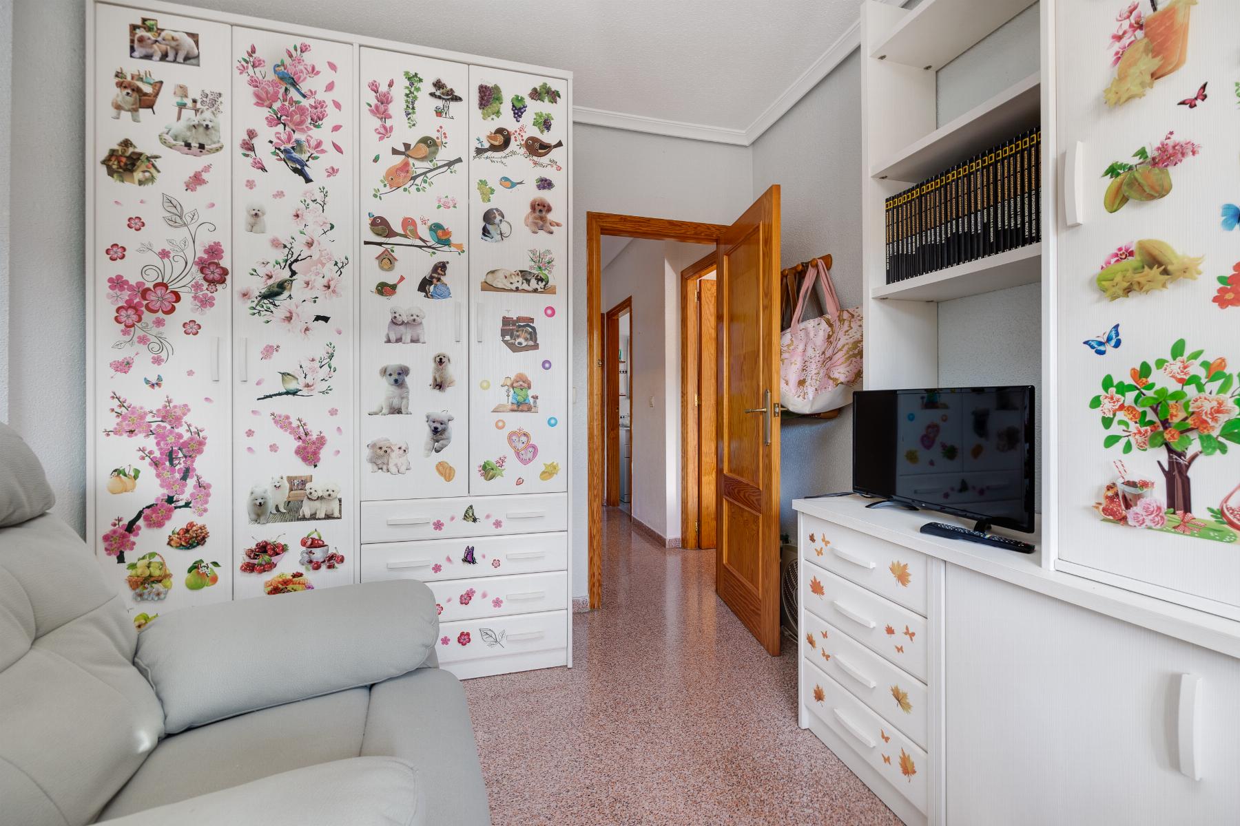 Living room corner with white storage cabinets covered in colorful dog and floral stickers, a gray sofa in foreground, and a TV on a white dresser.
