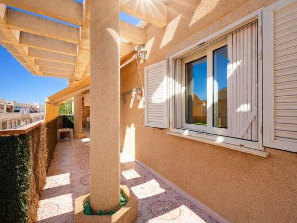 Sunlit beige balcony with a decorative tiled floor, white shuttered window, and a supporting column.