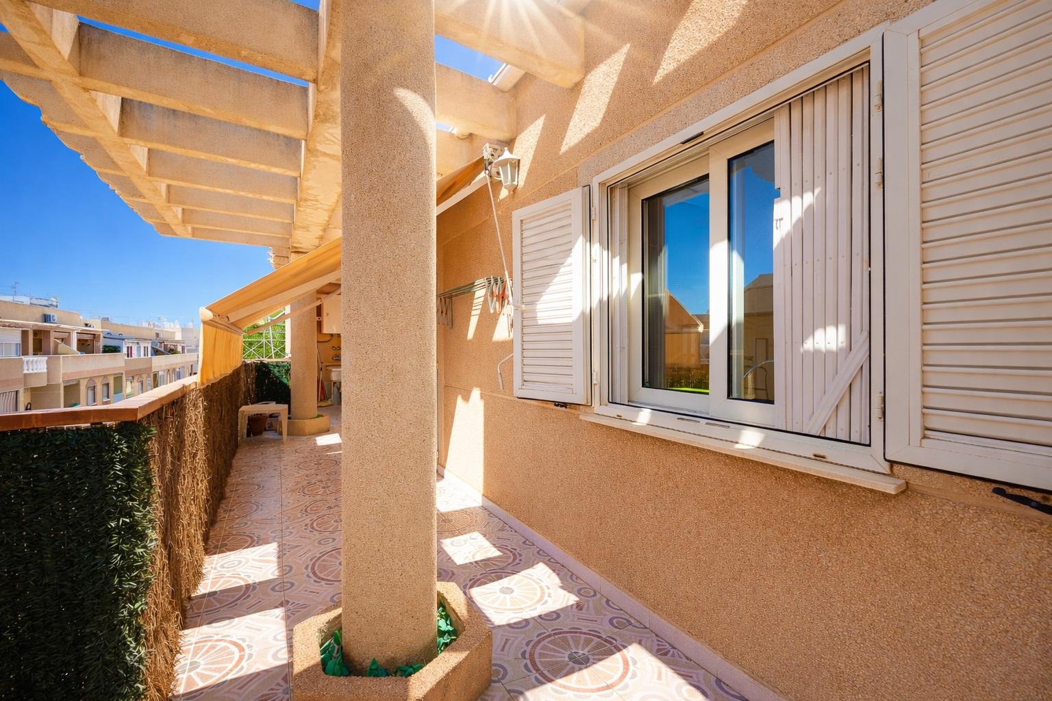 Sunlit beige balcony with a decorative tiled floor, white shuttered window, and a supporting column.