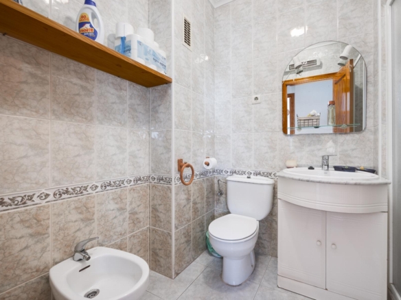 Small tiled bathroom with toilet, bidet, and white sink under a round mirror; wooden shelf with cleaning products on the wall.