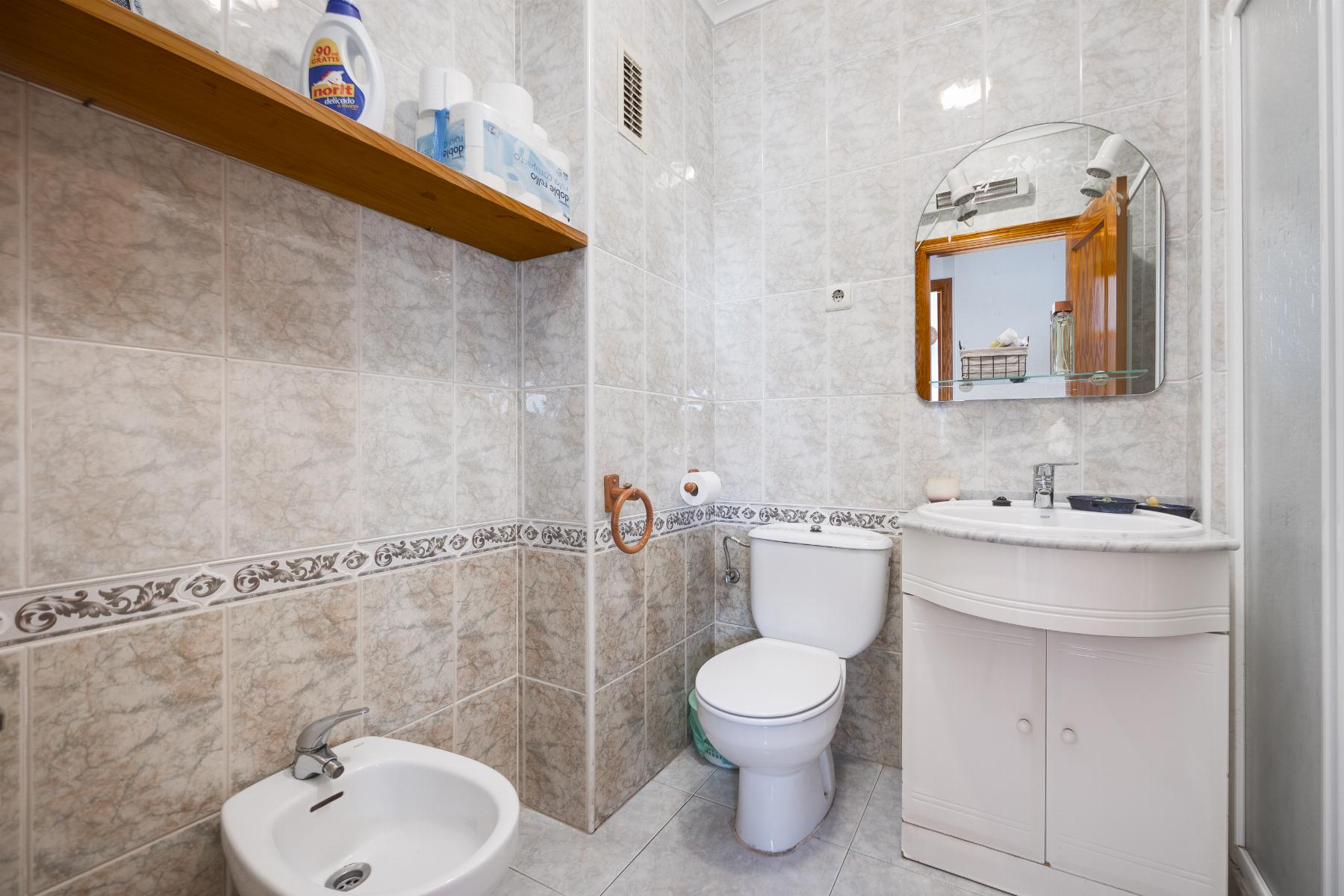 Small tiled bathroom with toilet, bidet, and white sink under a round mirror; wooden shelf with cleaning products on the wall.