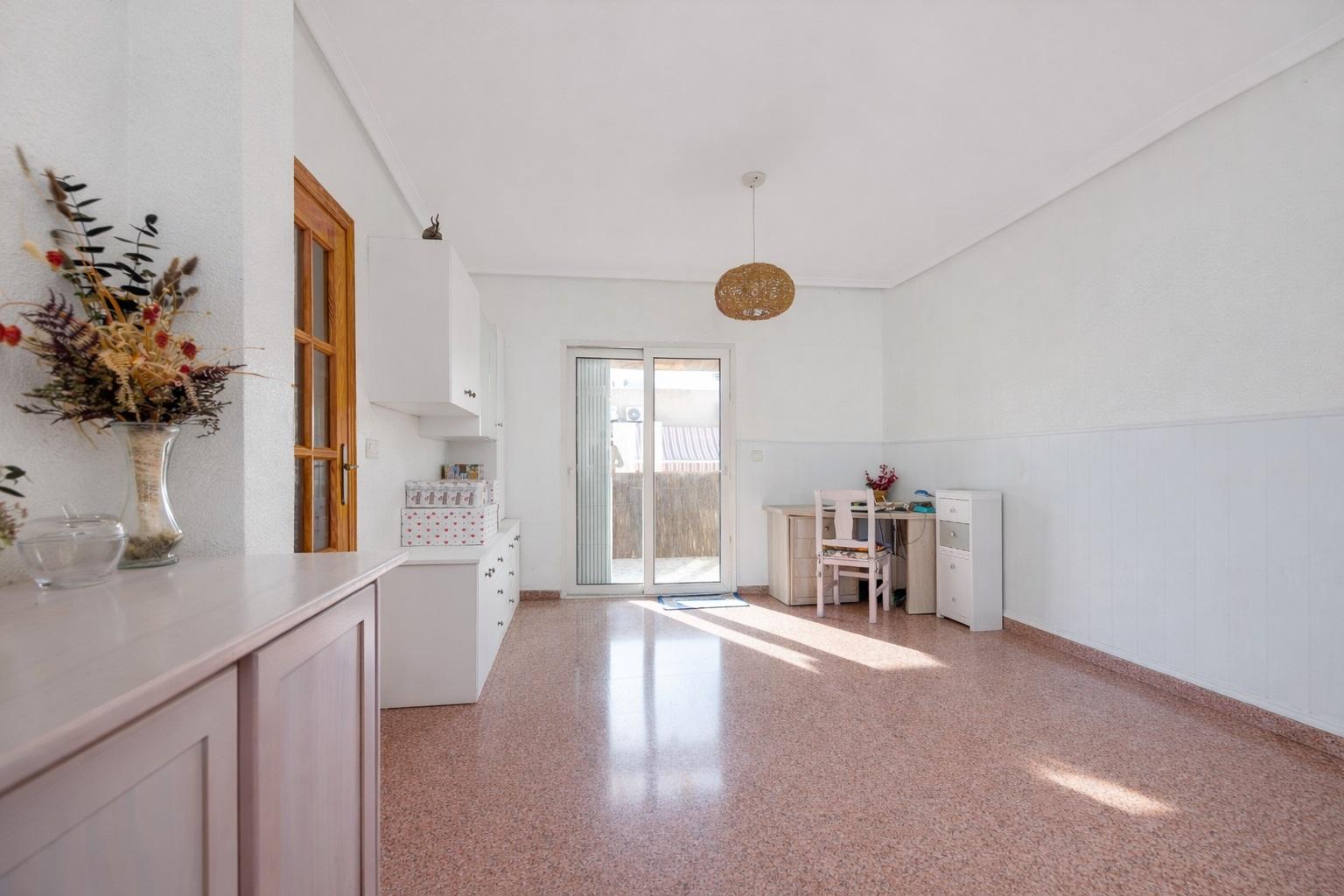Sunlit, minimalist room with white walls, pale furniture along the left wall, and a glass patio door at center.