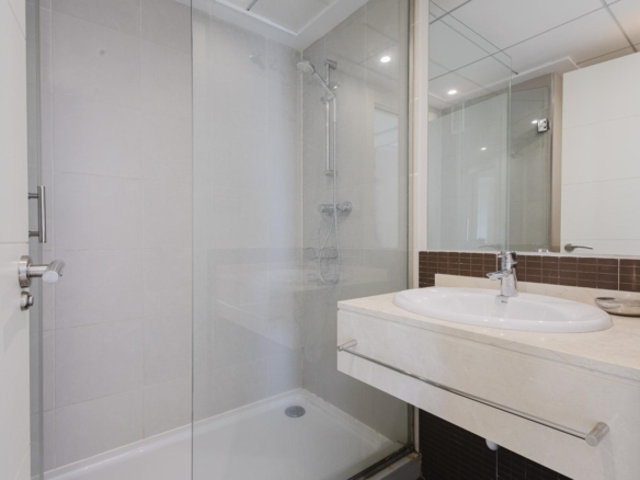 Modern bathroom with a glass enclosed shower, white sink, and marble countertop; brown tile backsplash and large mirror reflect recessed ceiling lights.