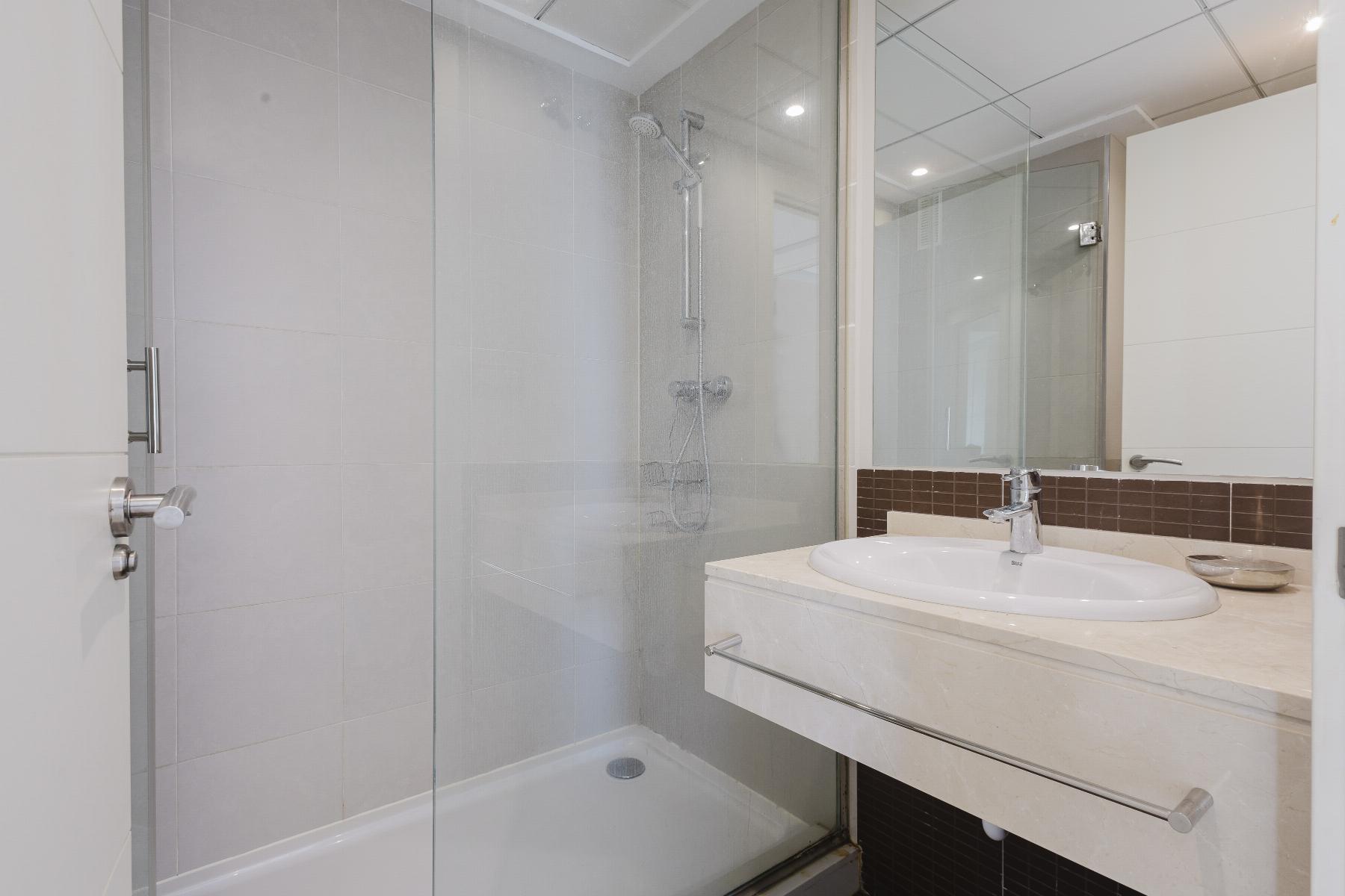 Modern bathroom with a glass enclosed shower, white sink, and marble countertop; brown tile backsplash and large mirror reflect recessed ceiling lights.