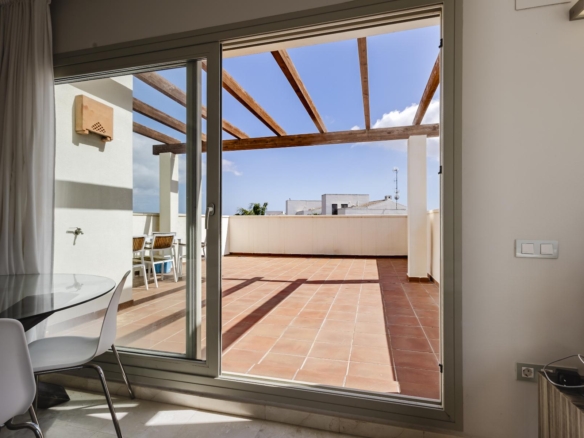 Sliding glass doors open to a terracotta-tiled terrace with wooden beams overhead and outdoor seating under a blue sky