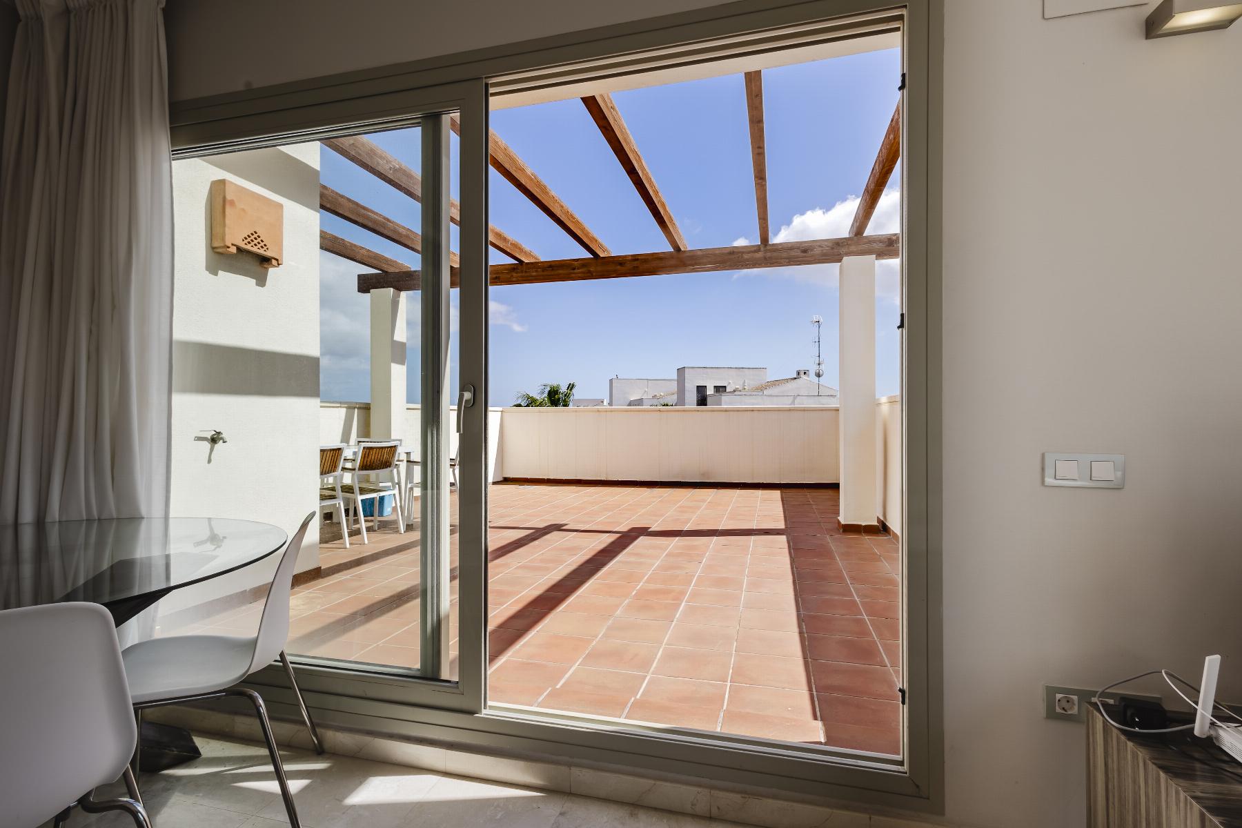 Sliding glass doors open to a terracotta-tiled terrace with wooden beams overhead and outdoor seating under a blue sky