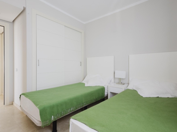 Two single beds with green throws in a bright, minimalist hotel room with a white headboard and bedside table between them.