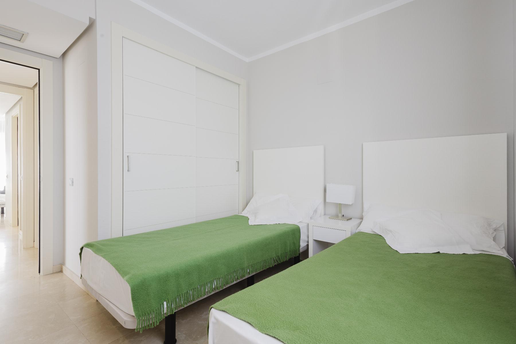 Two single beds with green throws in a bright, minimalist hotel room with a white headboard and bedside table between them.