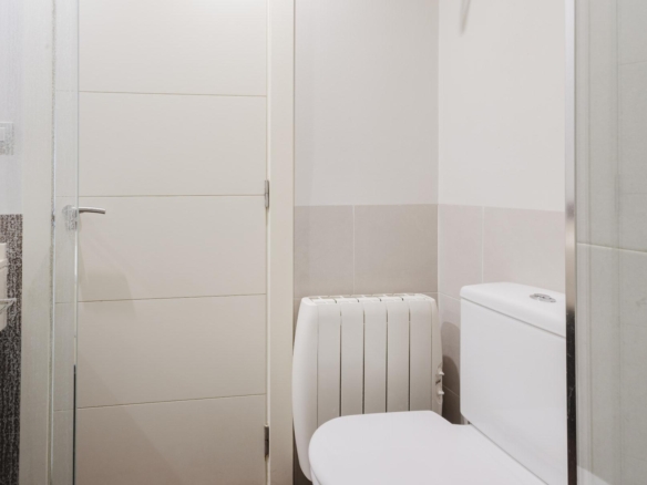 View of a small modern bathroom with a white toilet on the right, a vertical radiator in the middle, and light beige wall tiles near the door.