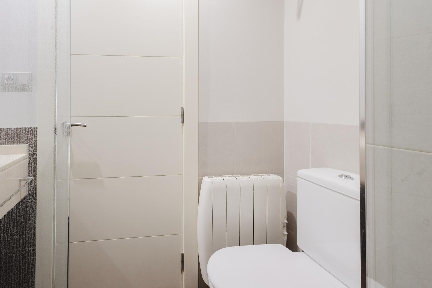 View of a small modern bathroom with a white toilet on the right, a vertical radiator in the middle, and light beige wall tiles near the door.