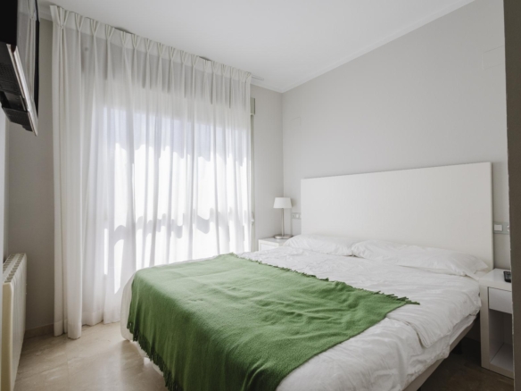 Minimalist hotel-style bedroom with a white headboard, bedding, green throw blanket, sheer white curtains, and a wall-mounted TV to the left.