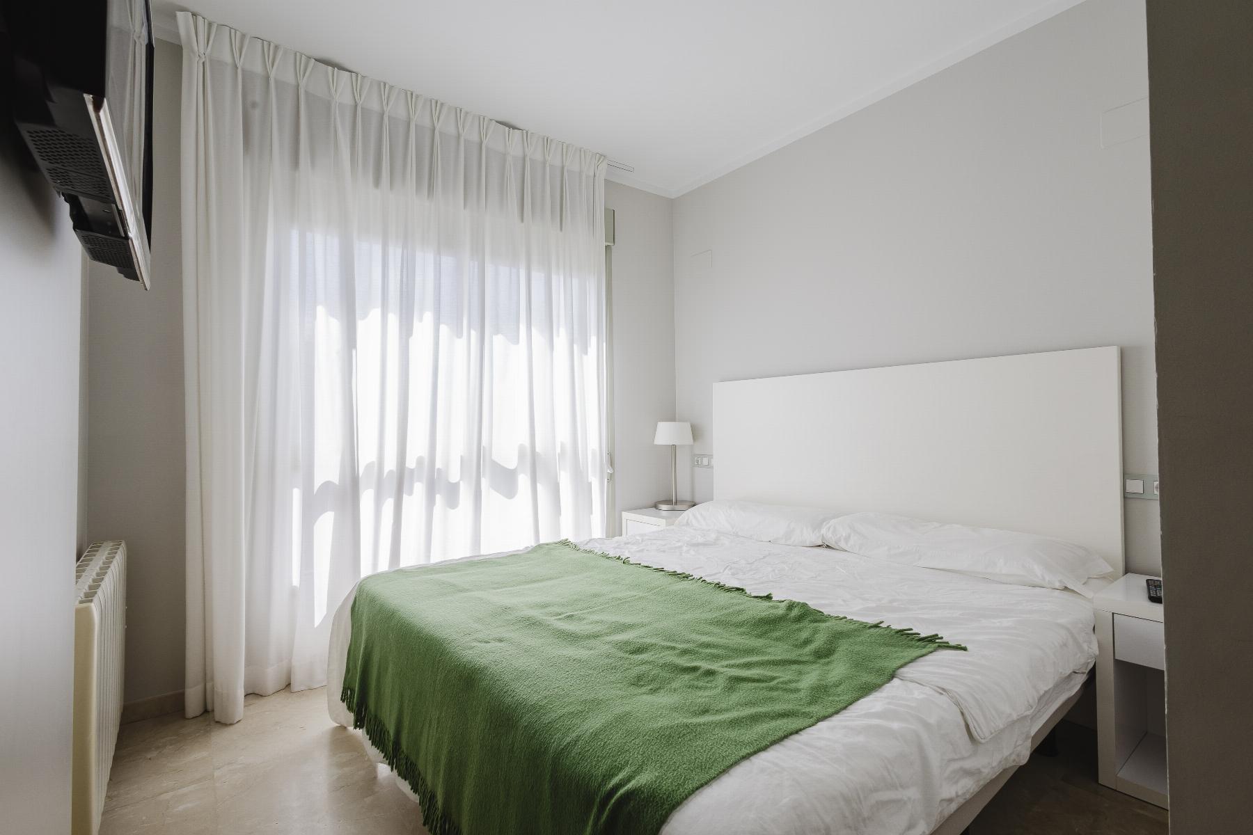 Minimalist hotel-style bedroom with a white headboard, bedding, green throw blanket, sheer white curtains, and a wall-mounted TV to the left.