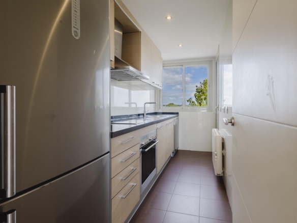 Narrow modern galley kitchen with a stainless steel fridge on the left, light wood cabinets, dark countertop, and a sink under a window at the far end showing blue sky and trees.