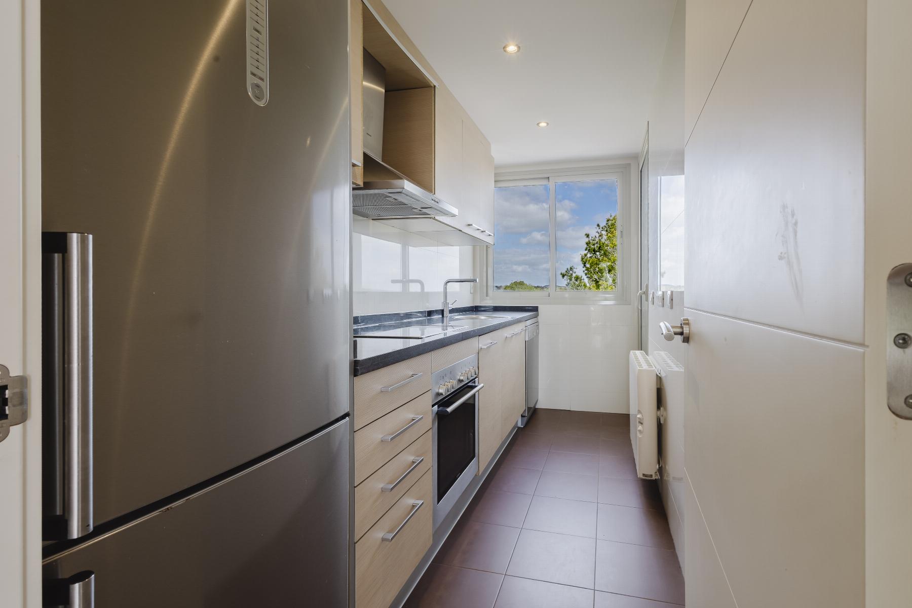 Narrow modern galley kitchen with a stainless steel fridge on the left, light wood cabinets, dark countertop, and a sink under a window at the far end showing blue sky and trees.