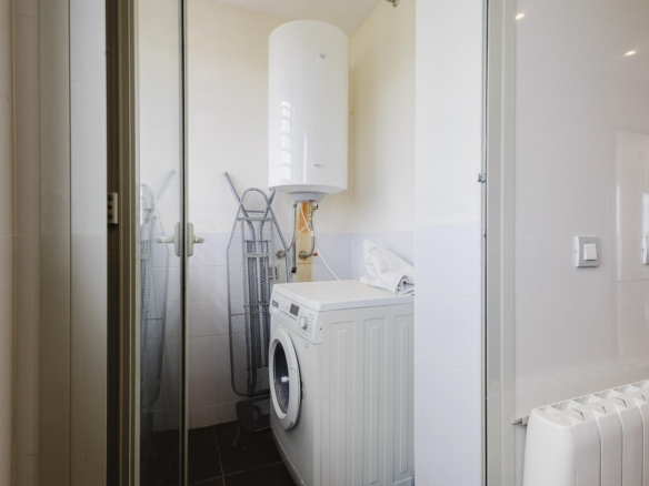 Compact laundry room with a front-loading washer, tall white water heater, and ironing boards nearby behind a glass door.
