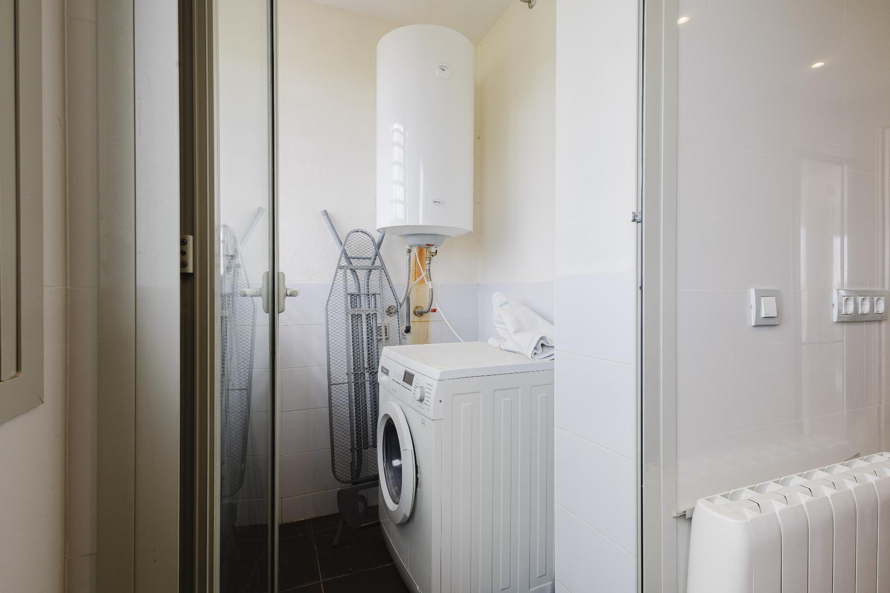 Compact laundry room with a front-loading washer, tall white water heater, and ironing boards nearby behind a glass door.
