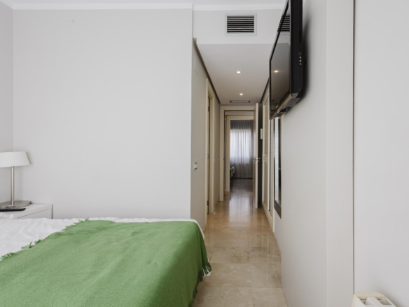 Hotel room with a bed in the foreground, green blanket, bedside lamp, and a hallway with a mounted TV on the right.