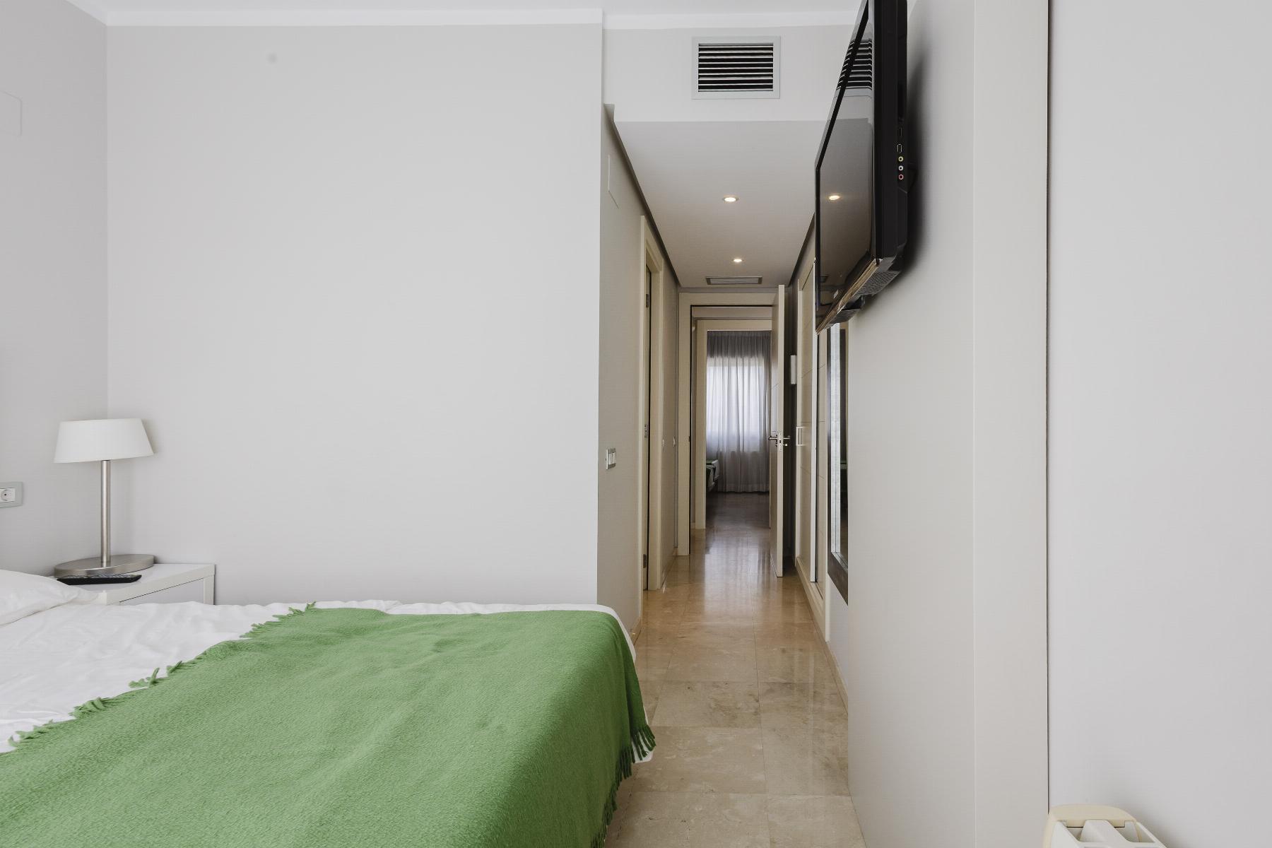 Hotel room with a bed in the foreground, green blanket, bedside lamp, and a hallway with a mounted TV on the right.