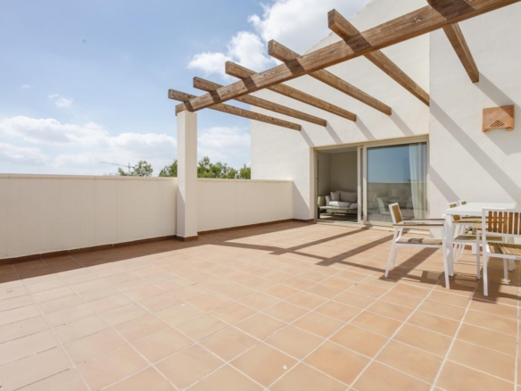 Terrace with tan tiled floor, white walls, and exposed wooden pergola beams; sliding glass doors open to a living room furniture set nearby under a sunny sky.