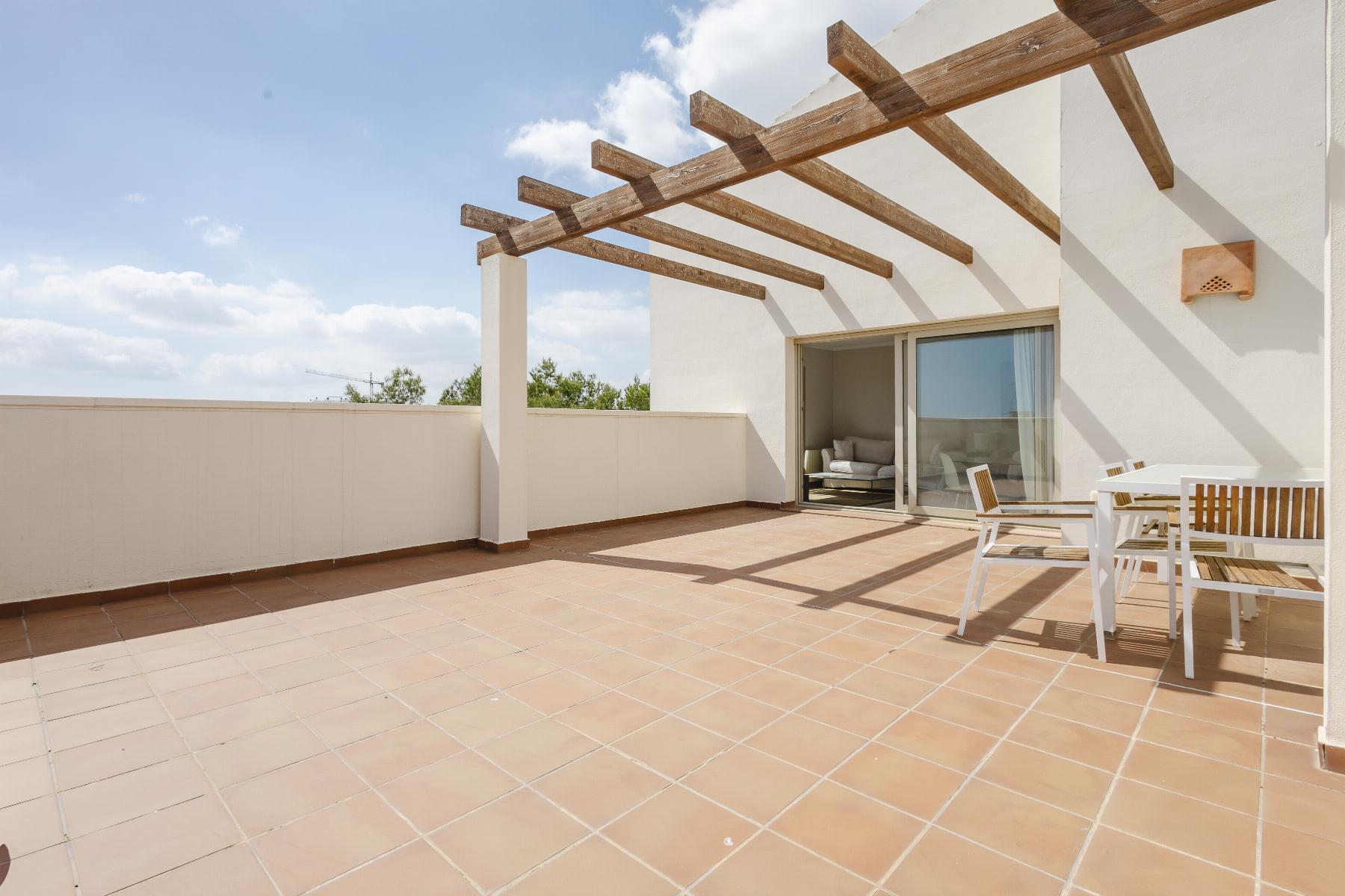 Terrace with tan tiled floor, white walls, and exposed wooden pergola beams; sliding glass doors open to a living room furniture set nearby under a sunny sky.