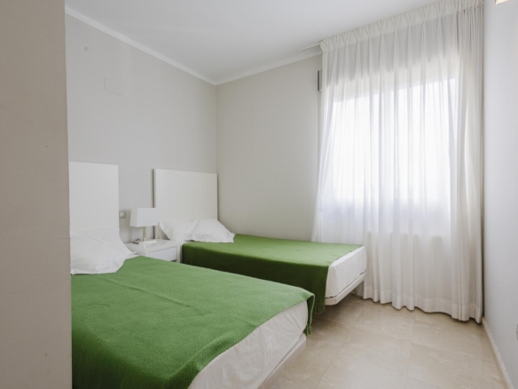 Twin hotel room with two single beds in green bedspreads and white headboards beside a nightstand and lamp under soft natural light.