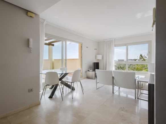 Bright open living/dining area with white seating, a glass dining table, and large windows/doors to a balcony with a light, neutral palette.