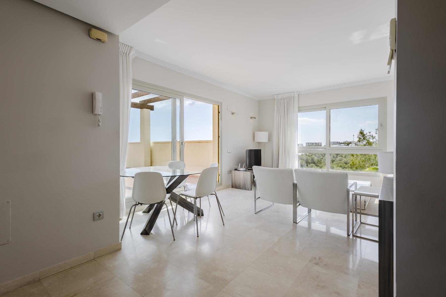 Bright open living/dining area with white seating, a glass dining table, and large windows/doors to a balcony with a light, neutral palette.