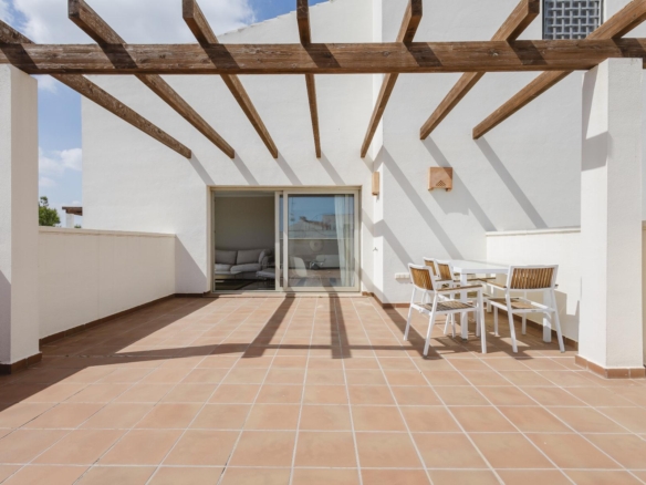 Terrace with white walls and a wooden pergola, sliding glass door to a living room, and a white table with wooden chairs on terracotta tiles.