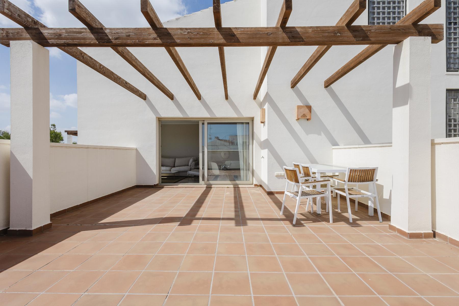 Terrace with white walls and a wooden pergola, sliding glass door to a living room, and a white table with wooden chairs on terracotta tiles.