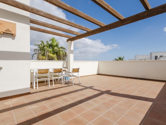 Terrace with terracotta tiles, white walls, and a wooden pergola; a white dining table with wooden chairs and palm trees under a blue sky in the background.