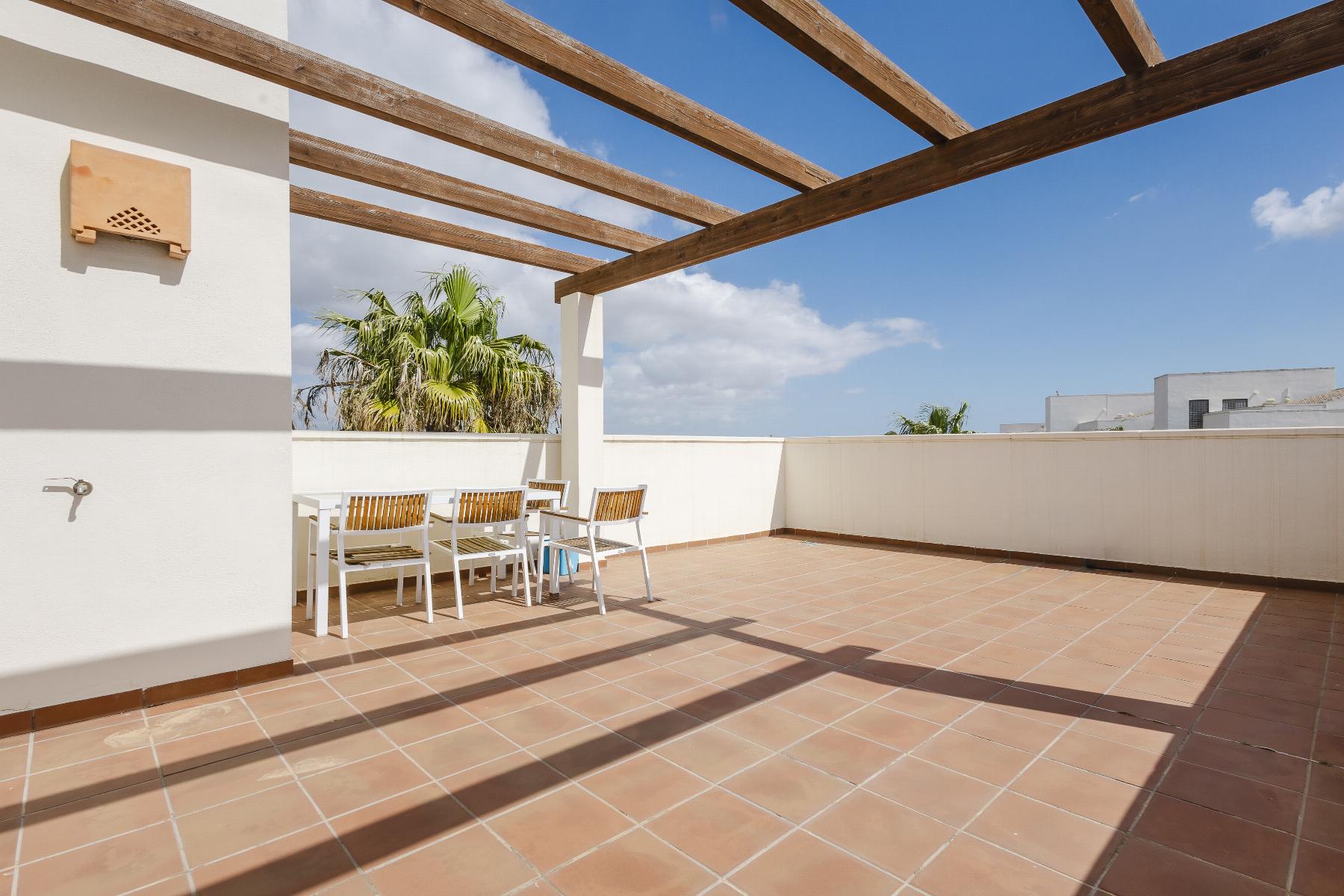 Terrace with terracotta tiles, white walls, and a wooden pergola; a white dining table with wooden chairs and palm trees under a blue sky in the background.
