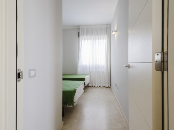 Narrow hallway opening into a bright bedroom with two single beds covered in green throws and a sheer white curtain window at the far end.