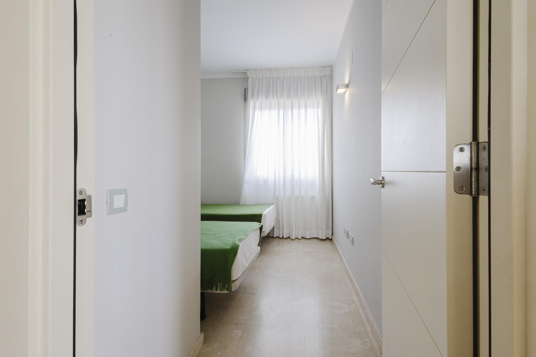 Narrow hallway opening into a bright bedroom with two single beds covered in green throws and a sheer white curtain window at the far end.