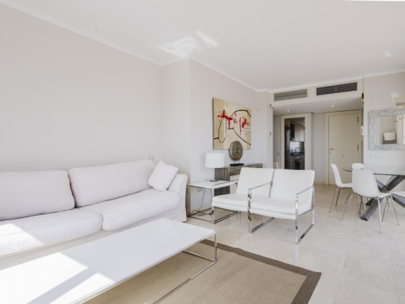 Bright, minimalist living room with a white sofa, white chairs, glass coffee table, and an abstract painting on the wall near the doorway to the kitchen area.