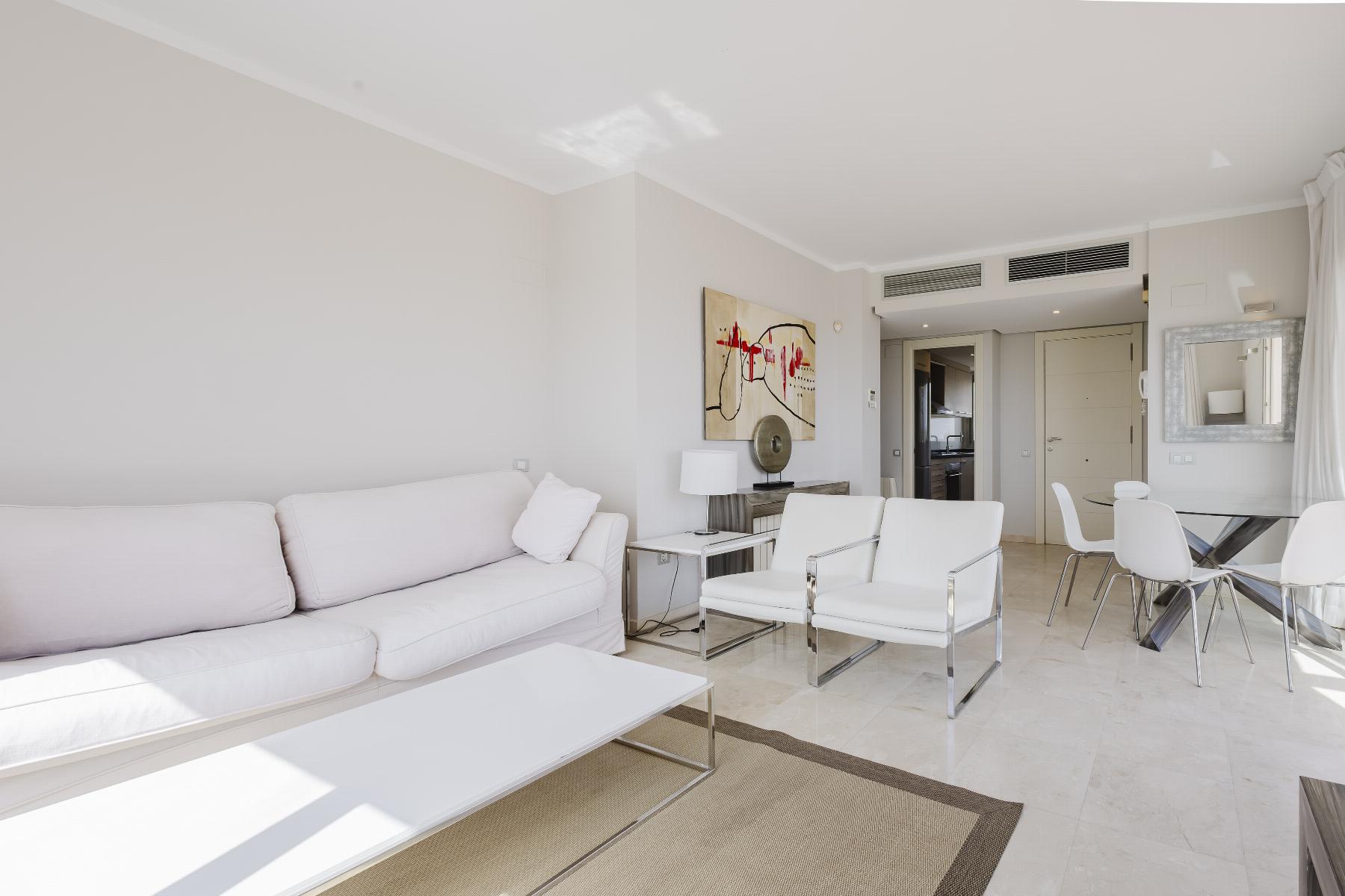 Bright, minimalist living room with a white sofa, white chairs, glass coffee table, and an abstract painting on the wall near the doorway to the kitchen area.