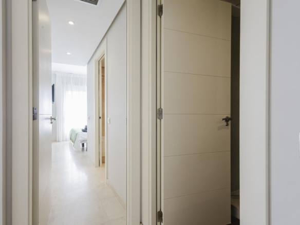 View down a bright white hallway with several doors, opening into a bedroom with a bed and sheer curtains