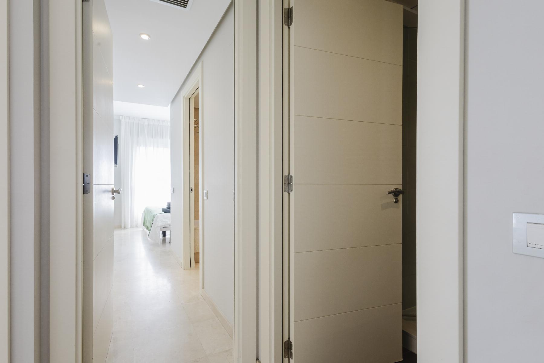 View down a bright white hallway with several doors, opening into a bedroom with a bed and sheer curtains